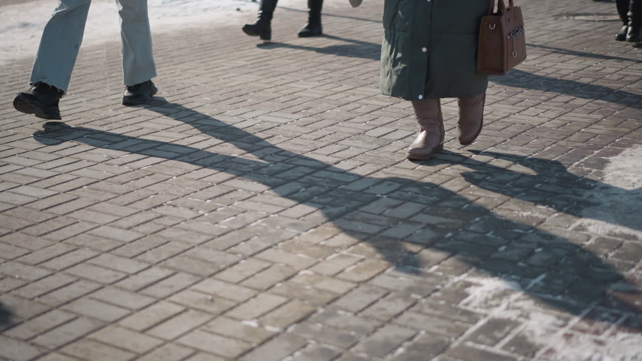leg level view of busy walkway as people in boots move along tiled pavement during rush hour, long shadows across bricks, patches of snow at edges, urban commute captured from low perspective
