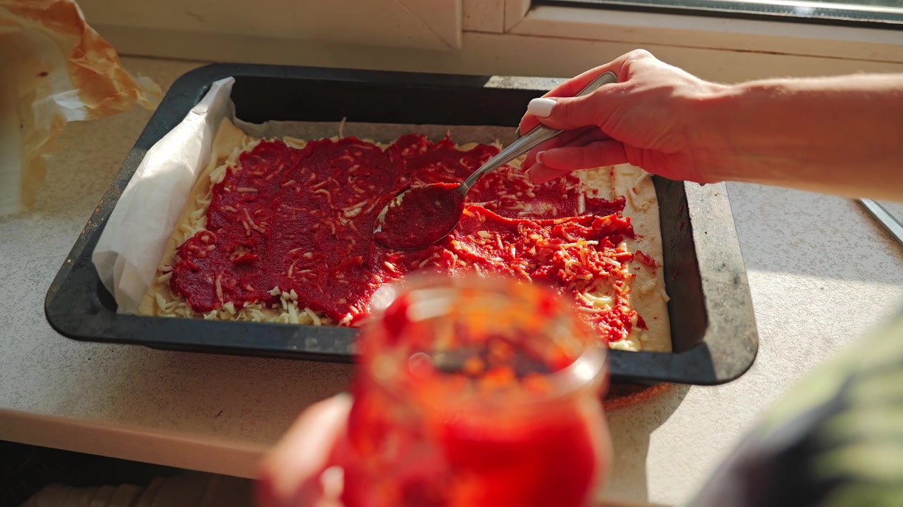 Hands carefully spread rich tomato sauce over shredded cheese before baking