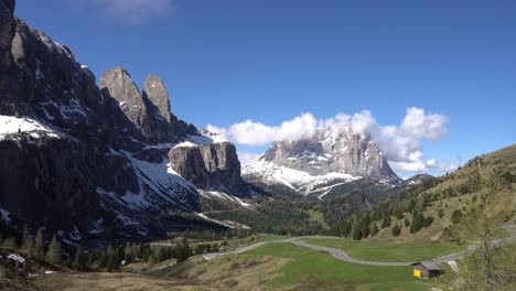 winding alpine road through green meadows and pine forest backdropped by snow-dusted peaks of the dolomites in italy. hyperlapse, panning shot