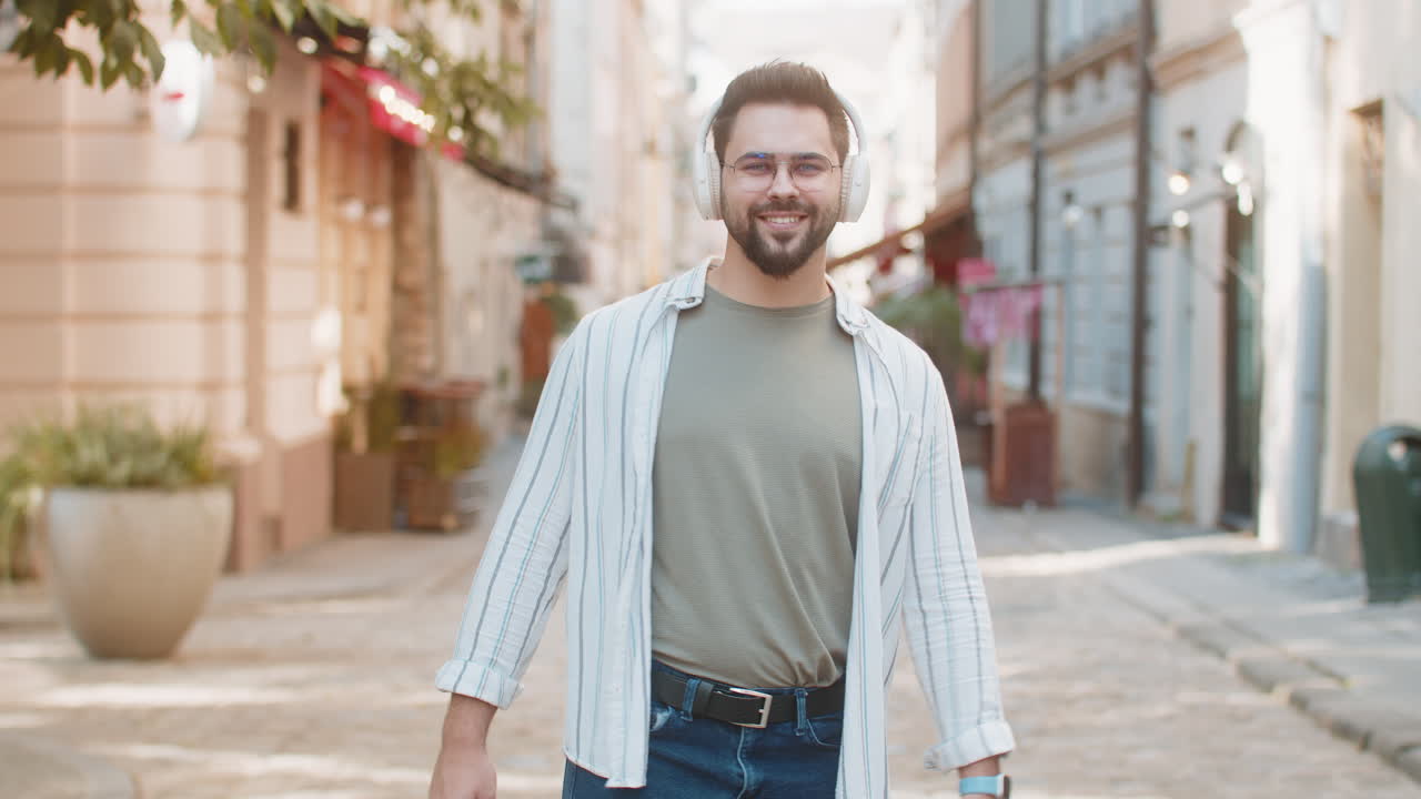 Happy young man in glasses listening music via wireless headphones while walking on city street