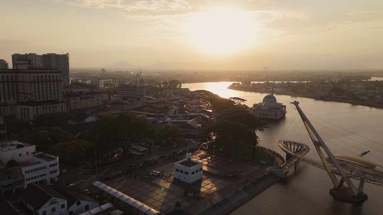 Aerial drone image of Kuching’s Darul Hana Bridge stretching over the Sarawak River during golden hour. The State Mosque and historic buildings surrounding the waterfront.