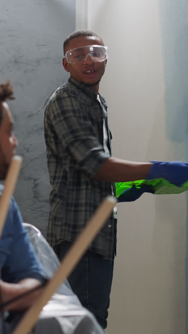 Cheerful African-American man paints wall talking to brother assembling chair in living room. Black guys repair home together. Apartment renovation