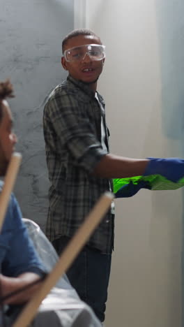 cheerful african-american man paints wall talking to brother assembling chair in living room. black guys repair home together. apartment renovation