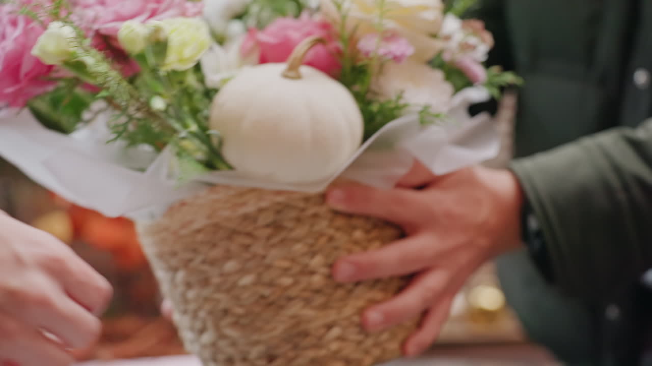 Primer plano de clavel rosa en un ramo. Manos de florista arreglando rosas y follaje en un mostrador rústico, colocando el arreglo envuelto en una cesta tejida. Tonos pastel suaves, movimiento delicado, ambiente acogedor de boutique.