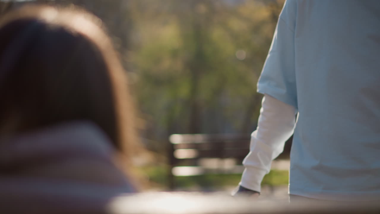 흰색 옷을 입은 남자가 앉아 있는 흰색 옷을 입은 여자 앞에서 배낭을 들고 있습니다. 손과 가방을 클로즈업했으며, 공원 벤치 배경은 부드럽게 흐릿하게 처리되었습니다. 무언가를 건네는 듯한 제스처, 햇빛이 비치는 테두리, 캐주얼한 교환이 세부적으로 포착되었습니다.