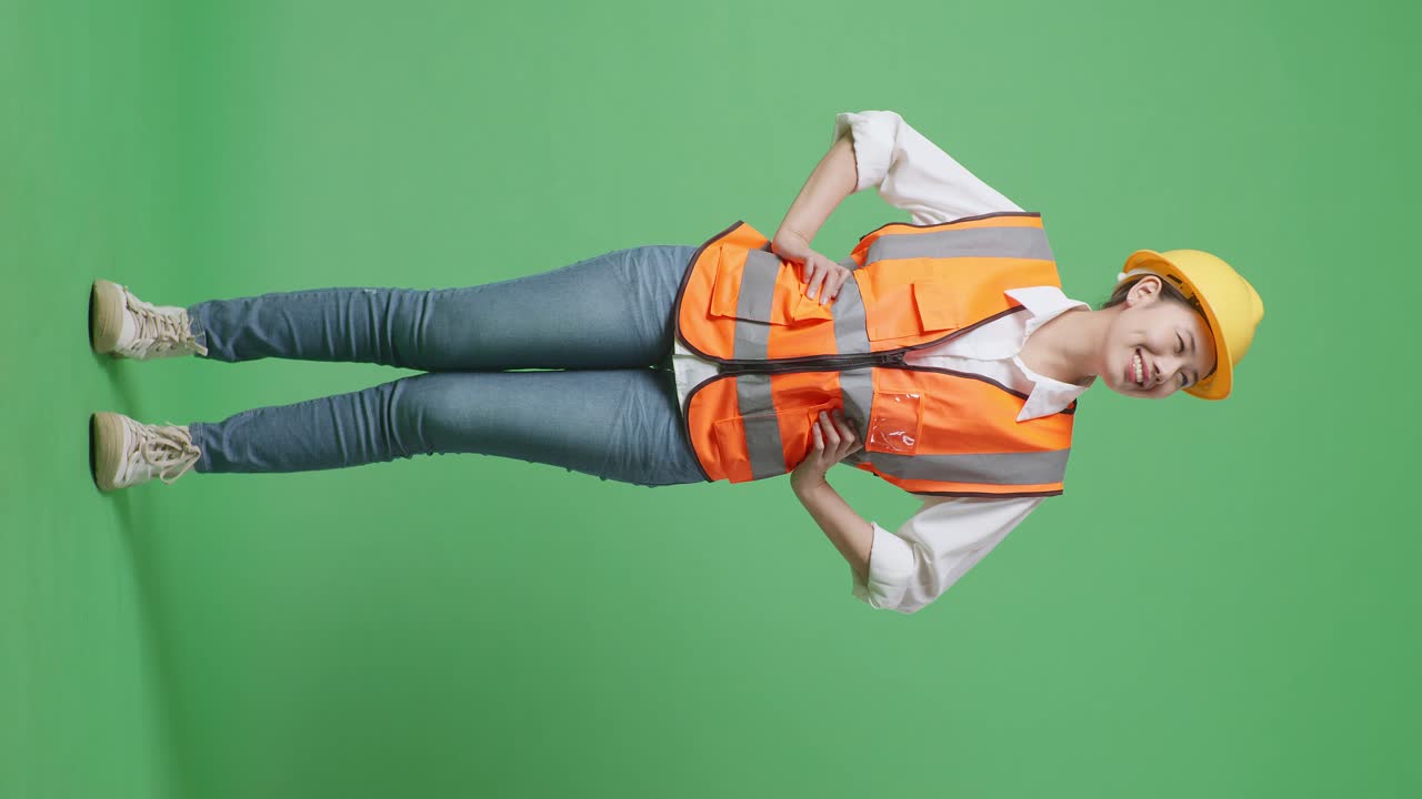 cuerpo lleno de ingeniera asiática que lleva casco de seguridad mirando a su alrededor mientras está de pie con los brazos akimbo en el estudio de fondo de pantalla verde