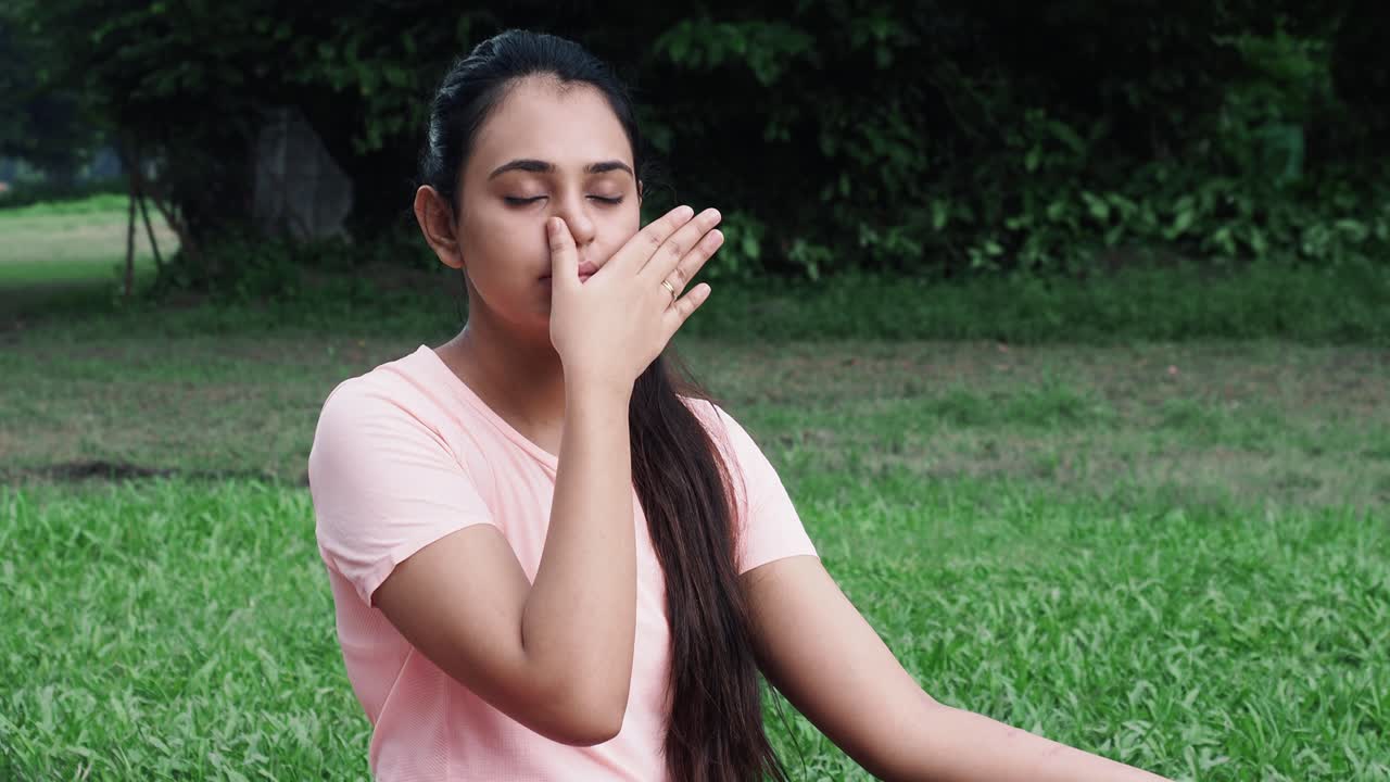 Medium shot of Young girl in sportswear doing nasal or nostril breathing exercise in park at kolkata - concept of self-care, yoga, workout and healthy lifestyle at indoors during coronavirus covid-19