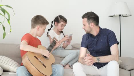 a teenager plays the guitar for his father and sister. a boy is learning to play the acoustic guitar. the family rests at home