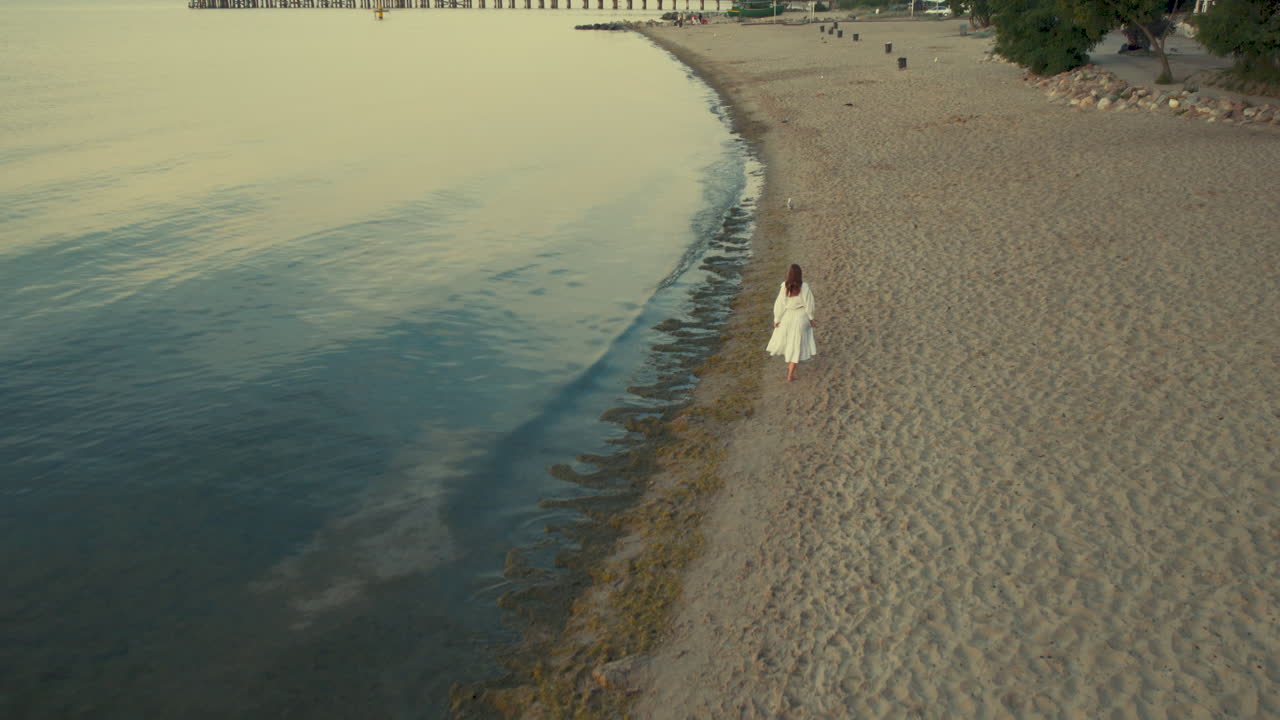 дрон следит за одинокой женщиной в белом летнем платье, идущей по пляжу на восходе солнца - задний вид