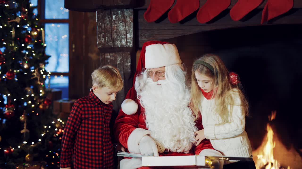 Santa Claus in a traditional red suit sitting in the middle of the room and telling stories for the boy and girl with the images in the album, which he is holding in his hands near the fireplace on a cold winter evening.
