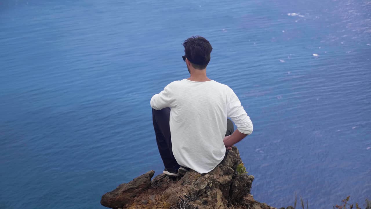 A man sitting on a rocky cliff and overlooking the beautiful blue waters of Madeira, Portugal - Wide shot