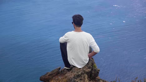 a man sitting on a rocky cliff and overlooking the beautiful blue waters of madeira, portugal - wide shot