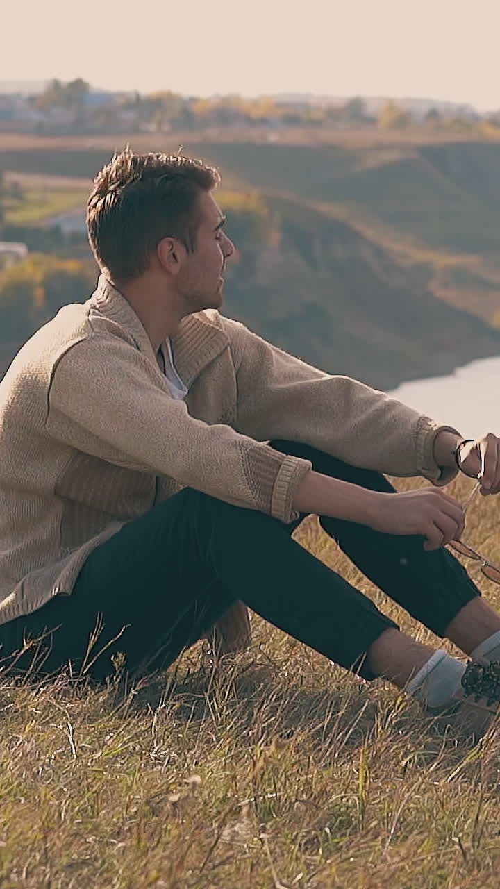 frustrated guy in jeans and pullover with hangover sits on river bank and holds grass stem in hand slow motion