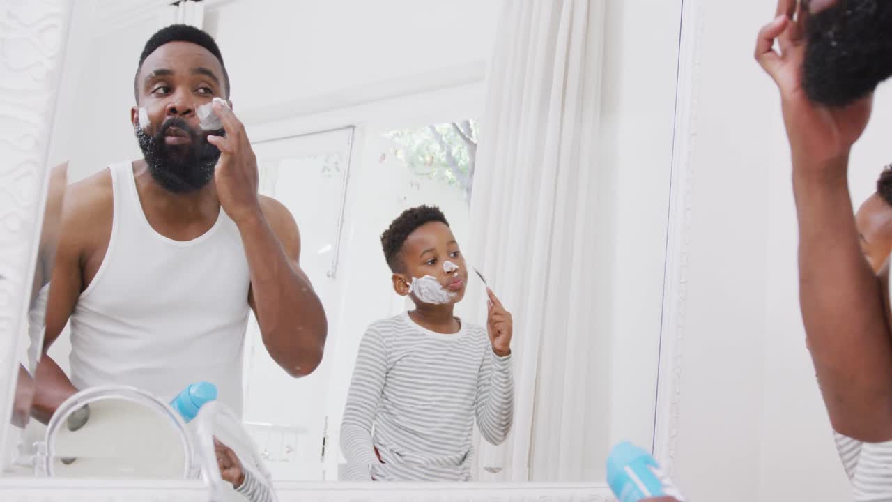 Happy african american father and son applying shaving cream on face in bathroom, in slow motion