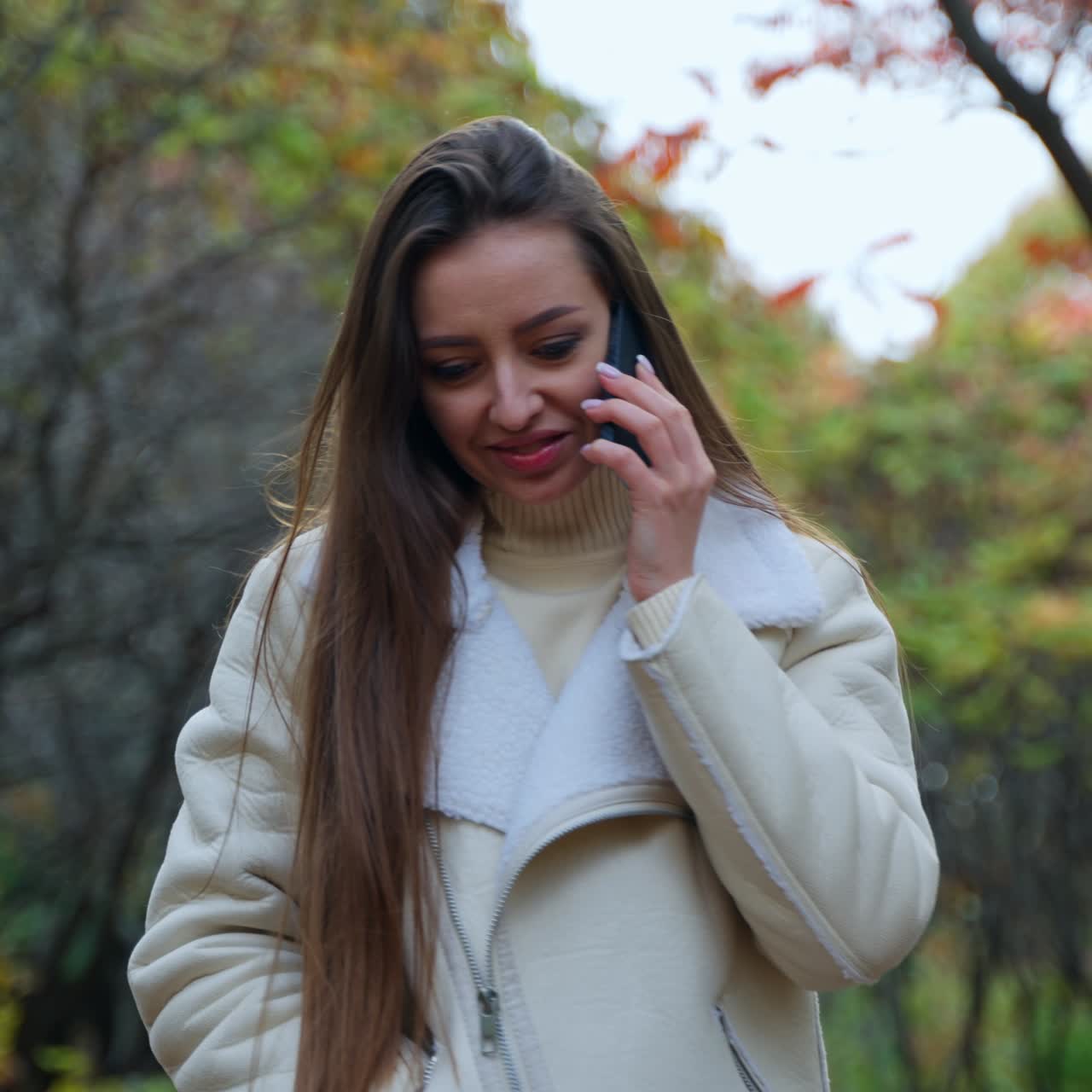 Caucasian lady speaks on the phone walking by the autumn nature. Woman touches her long hair constantly and smiling at conversation