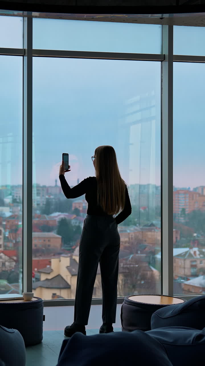 Young woman with a phone near window. Panoramic office view with large windows on city background. Rear view of a girl indoors photographing the evening city landscape. Vertical video