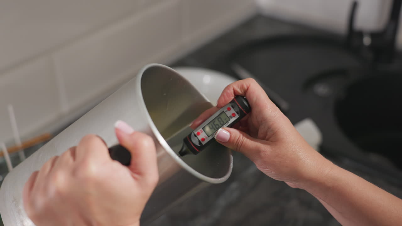 Hand view of cake baker using digital thermometer to monitor temperature of heated baking butter inside metal container during preparation in modern kitchen setup with precise attention to detail