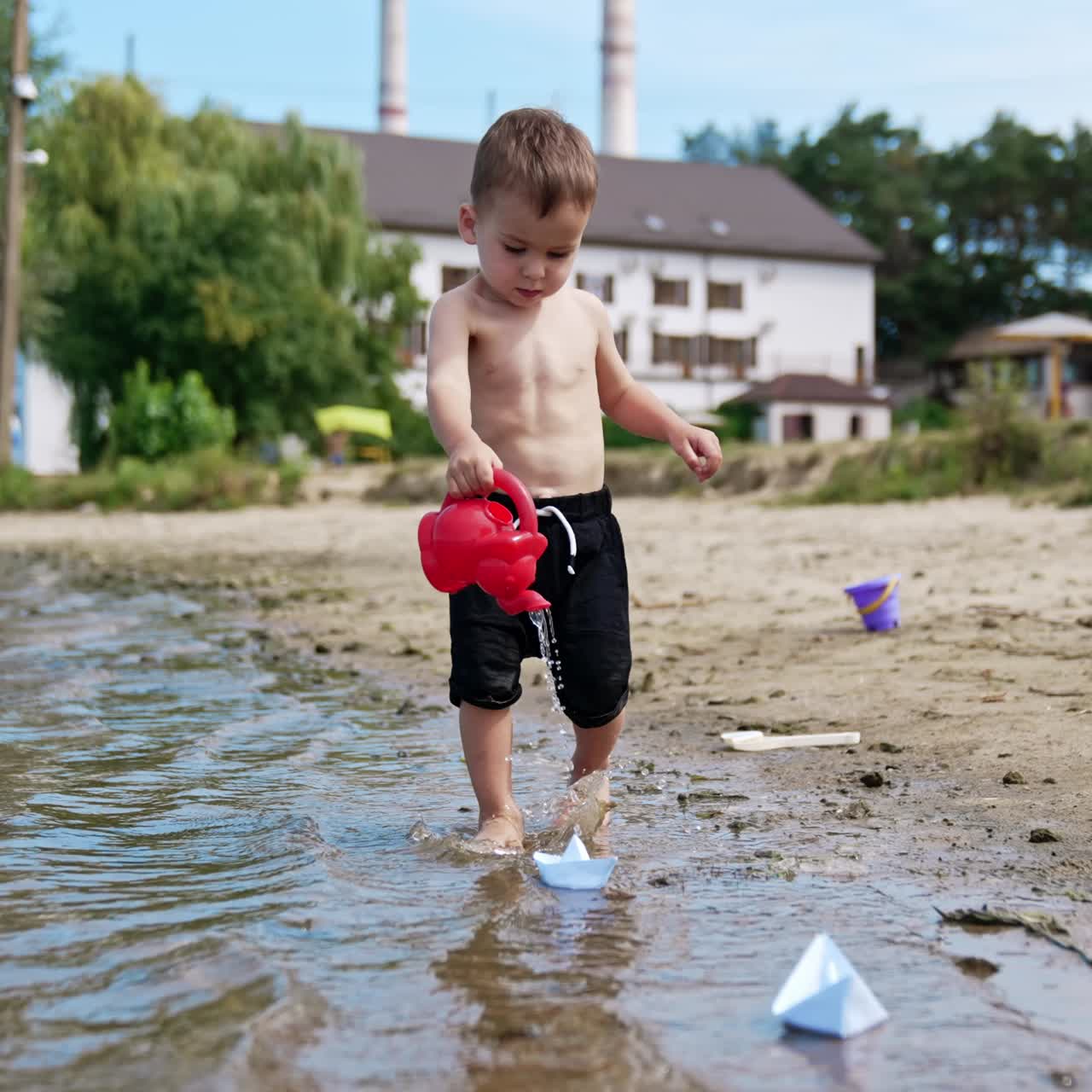 Adorable baby boy with a red watering can pouring water. Sweet child playing on the shore in summer