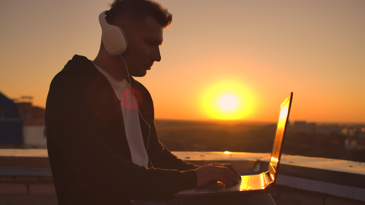 Uomo di spalle con le cuffie che ascolta musica e lavora sul tetto di un edificio al tramonto, con vista sulla città dall'alto. Tetto di un grattacielo al tramonto.