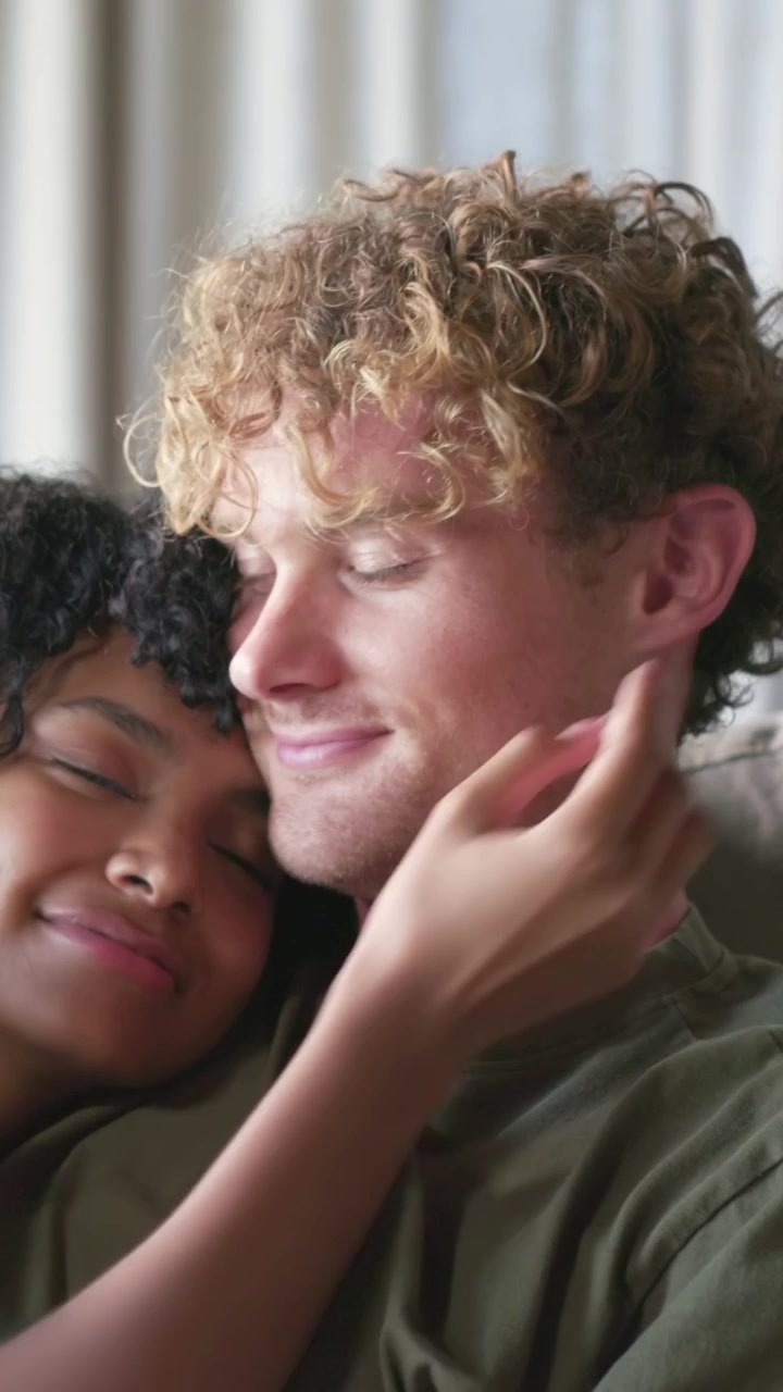 Vertical video: Leaning seated couple smiling at offscreen cue, wrapping arm, cupping face on couch