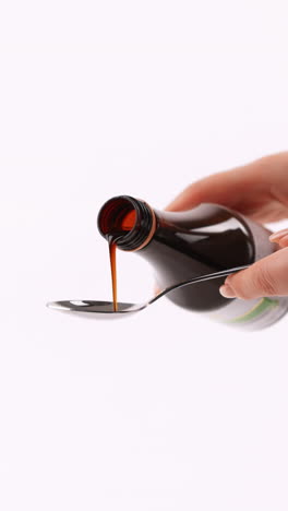 vertiendo jarabe de medicina en una cuchara de una botella marrón, sostenida por las manos de una mujer sobre un fondo blanco