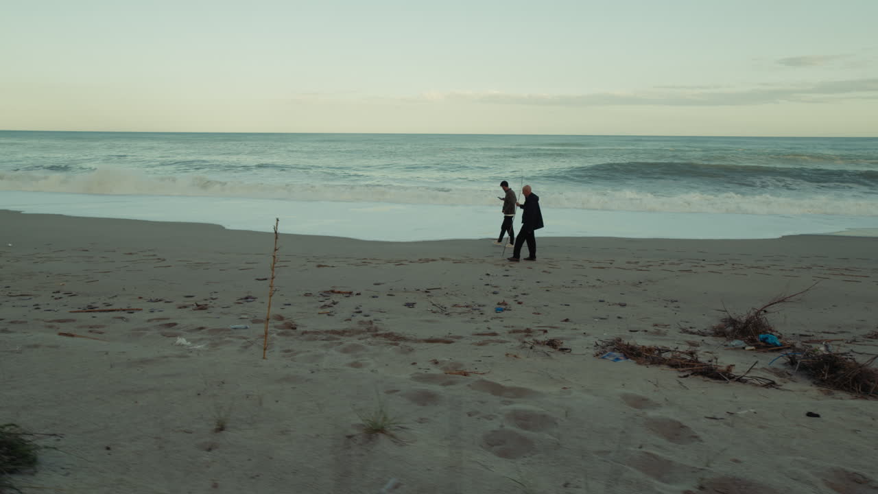 On A Long Beach, Two People Walk While Watching The Fury Of The Ocean Waves