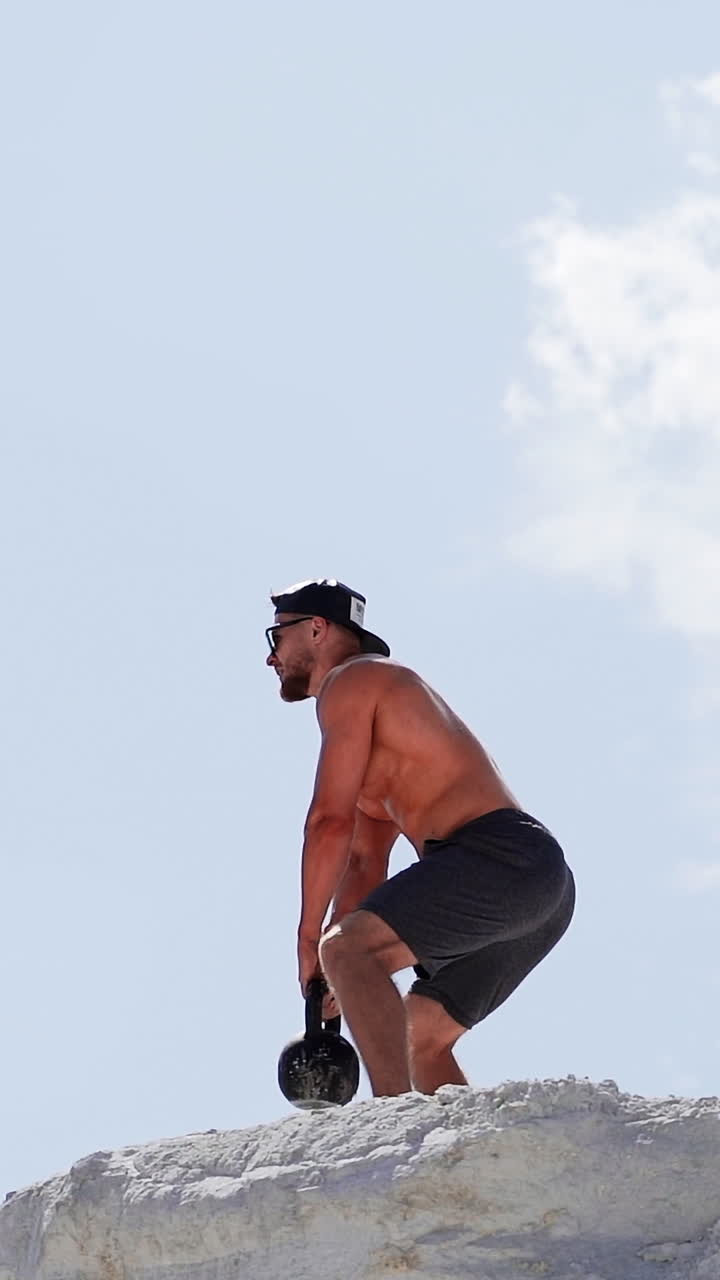 Side view of a strong sportsman training with heavy weight outside. Muscular athlete swings kettlebell on the high hill in a daytime. Vertical video