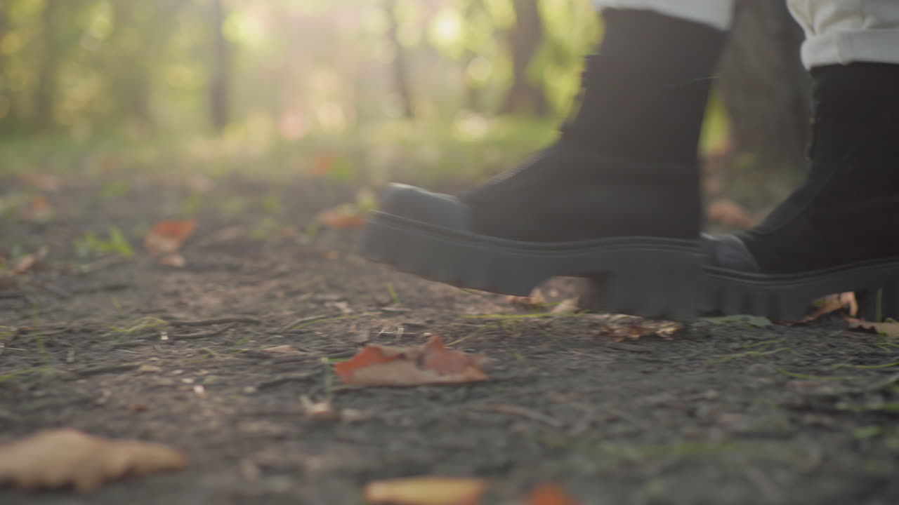 Side leg view of wanderer in boots strolling leisurely along forest path, heel lifting from ground, crisp leaves and soil under steps, soft sunlight and blur background suggesting calm autumn walk