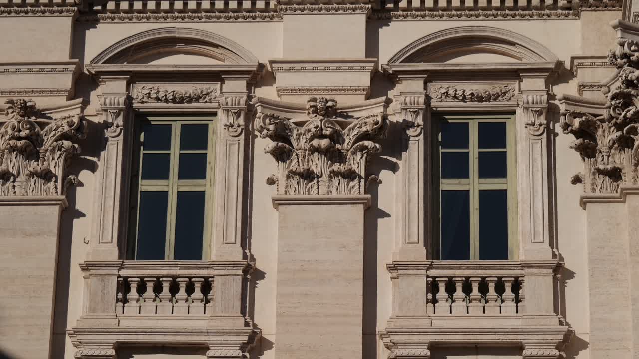 Close-up of a historic baroque façade in Rome, Italy, featuring symmetrical windows, decorative stone carvings, and Corinthian capitals illuminated by sunlight.