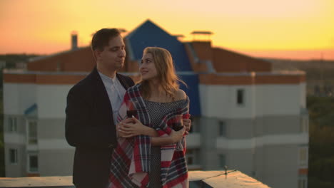 young married couple on the roof hugging and drinking red wine from glasses standing dressed in plaid and admiring the beautiful sunset over the city..