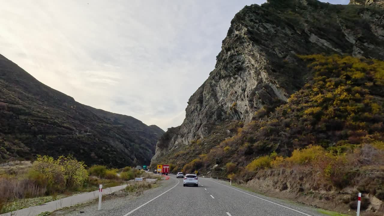 Cars navigate winding roads amidst Queenstown's rugged mountains. Clear skies and natural beauty enhance the serene drive