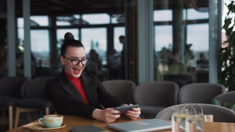 una joven alegre y hermosa con aparatos ortopédicos se sienta en un elegante restaurante con ropa de negocios, jugando felizmente a un juego en su teléfono inteligente
