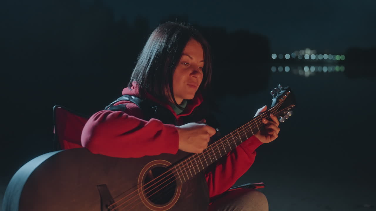 Musicista donna si gode una tranquilla serata al lago. Donna serena si esercita con la chitarra accanto a un lago tranquillo. Scena notturna calma con una donna che strimpella la chitarra accanto a un falò ardente vicino al lago.