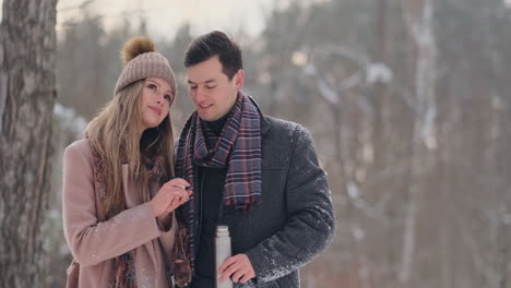 happy couple holding hot tea cups over winter landscape. young couple in love on a winter vacation, standing next to a tree and drinking hot cup of tea