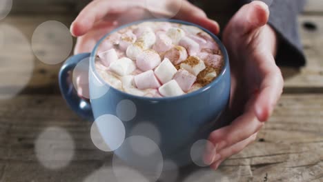 animation of light spots over hands holding mug with cocoa on wooden background