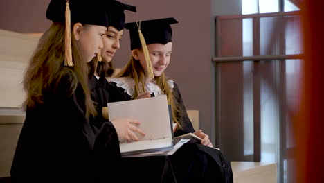 Vista-Lateral-De-Tres-Niñas-Felices-Con-Toga-Y-Birrete-Hablando-Y-Mirando-Sus-Cuadernos-Mientras-Se-Sientan-En-Las-Escaleras-En-La-Ceremonia-De-Graduación-Preescolar