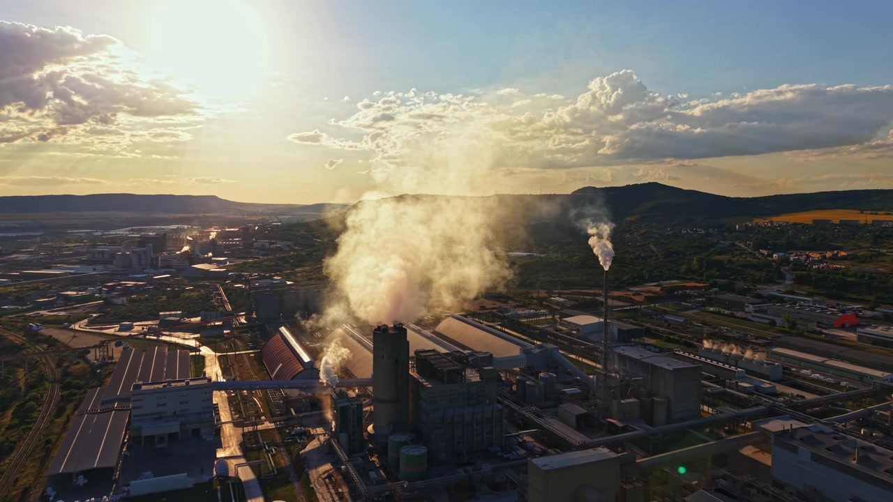 Fumo si alza da edifici industriali al tramonto su un paesaggio urbano, mostrando fabbriche e aree di produzione.