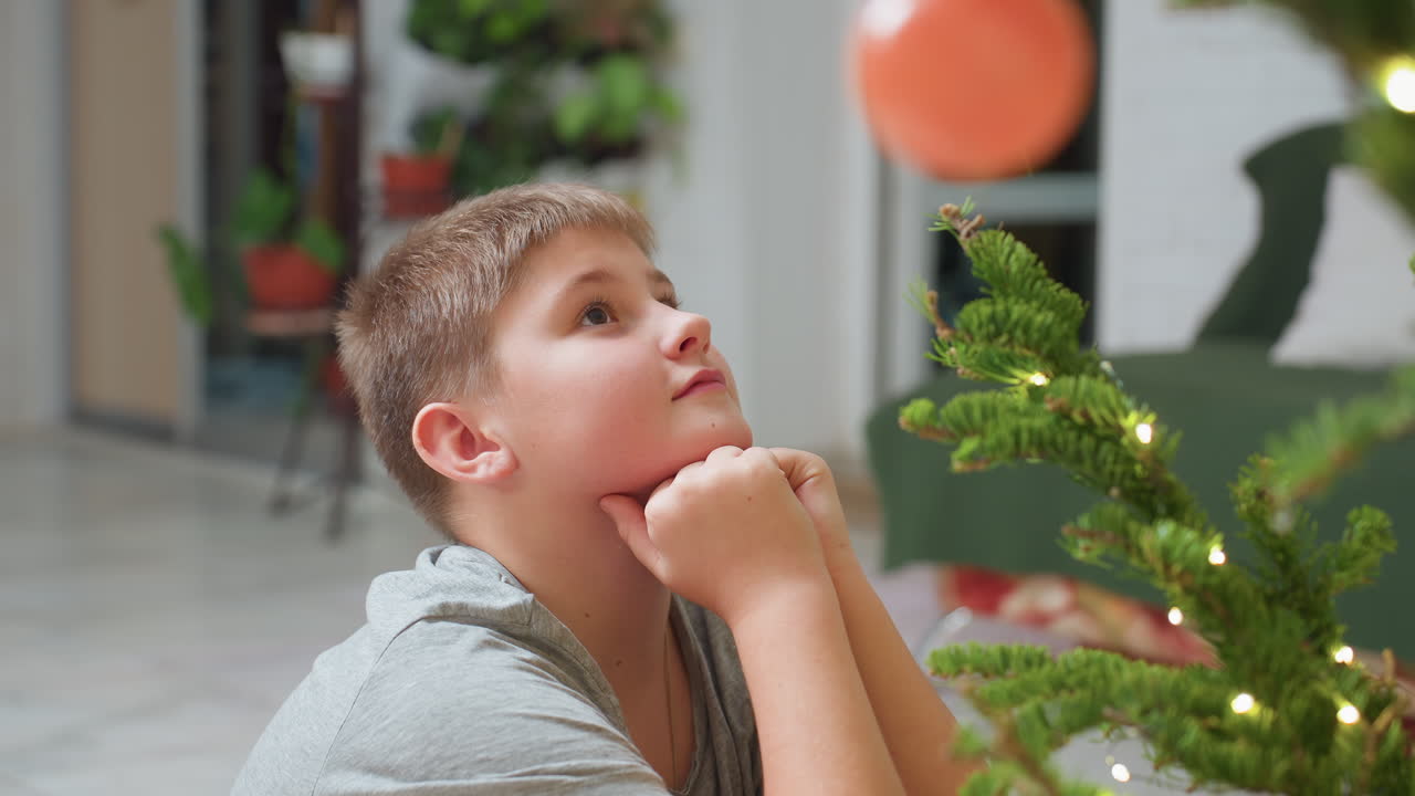 Boy sitting with hands under chin looking at decorated christmas tree, wearing grey top, peaceful indoor setting, thoughtful expression, cozy atmosphere surrounded by warm festive lights