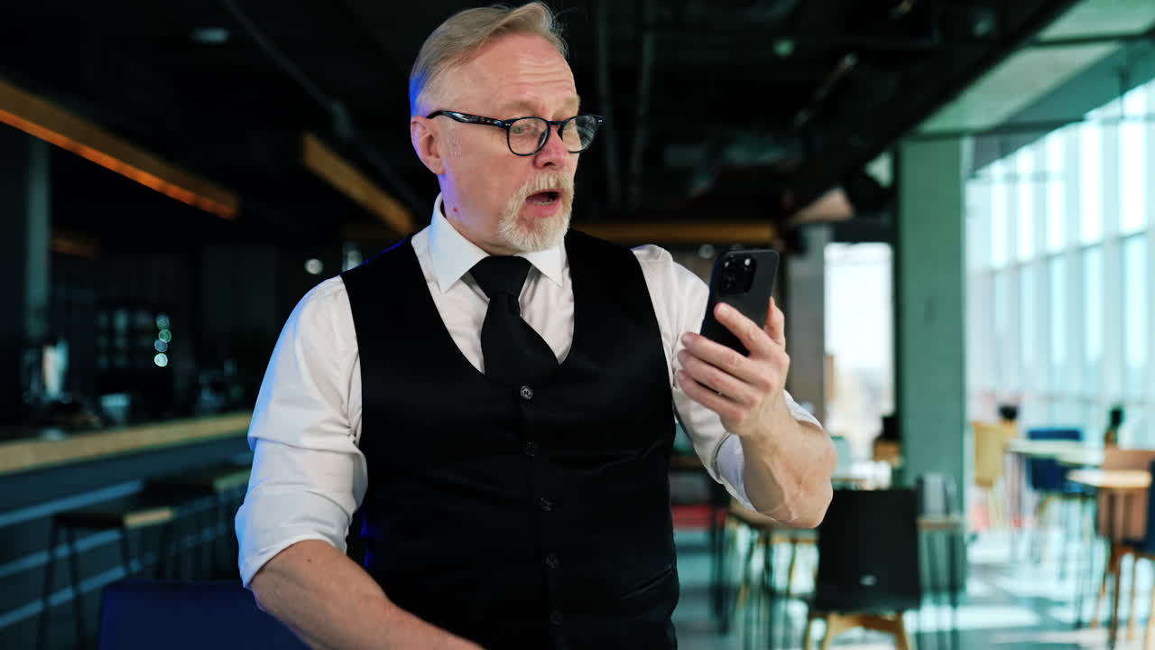 Mature man wearing white shirt, black vest, tie and glasses speaks on the phone. Man having a video chat and gestures actively.