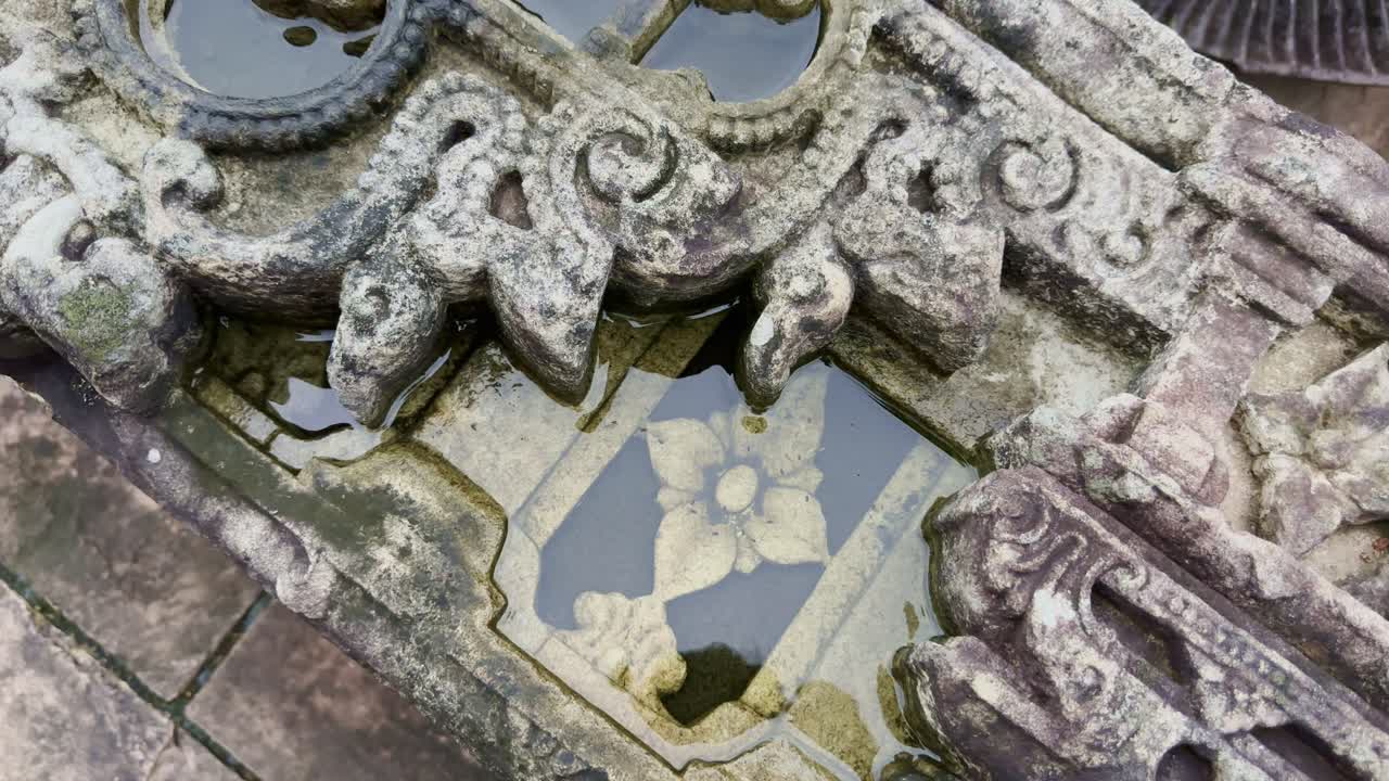 The camera spins around a weathered carved stone fragment, its surface adorned with intricate scrolls and a central floral motif, softened by age and rainwater collecting in its grooves