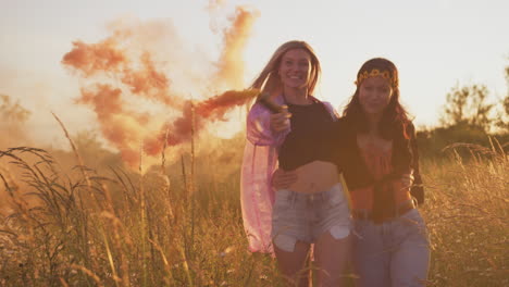 two female friends camping at music festival running through field with smoke flares