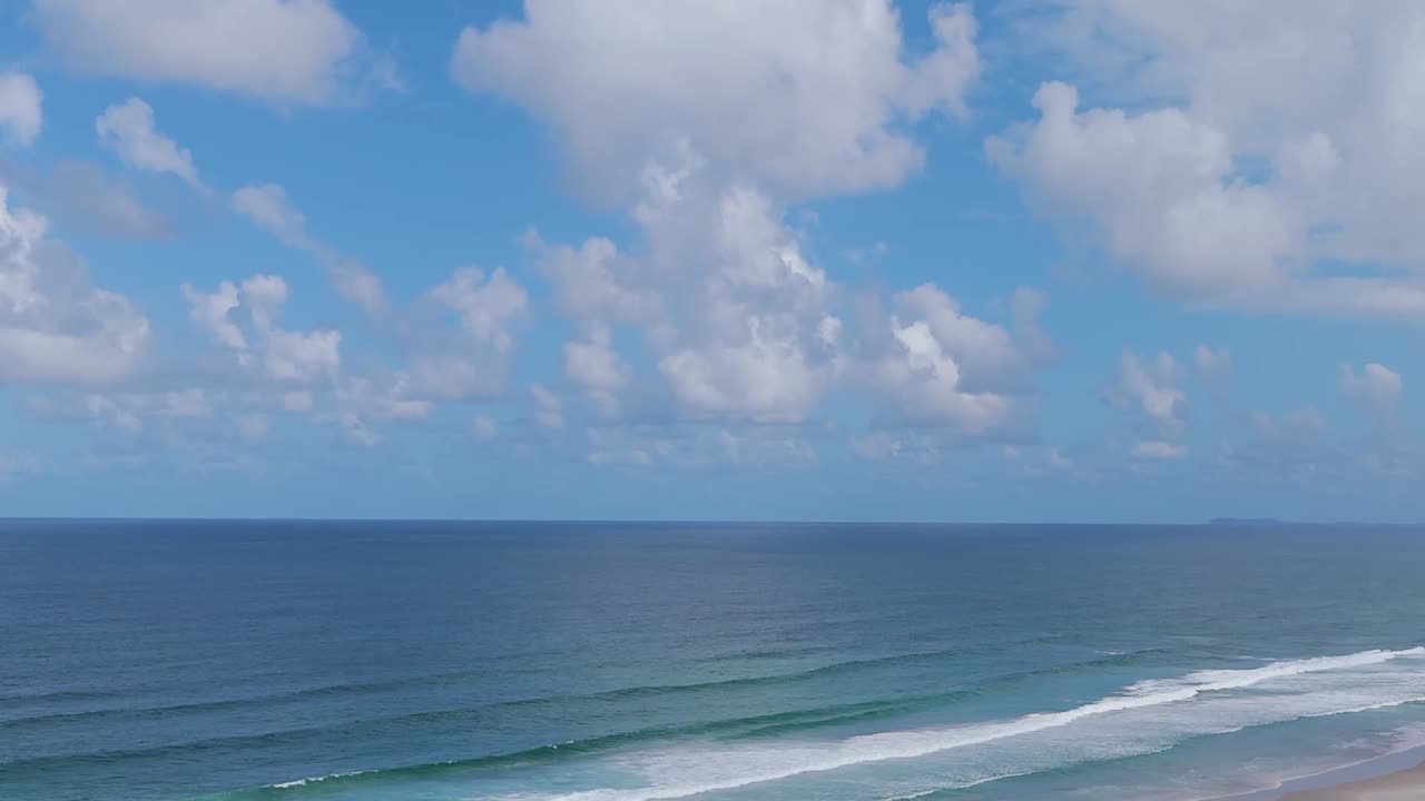Aerial view of ocean waves rolling towards the shore beneath a partly cloudy sky.