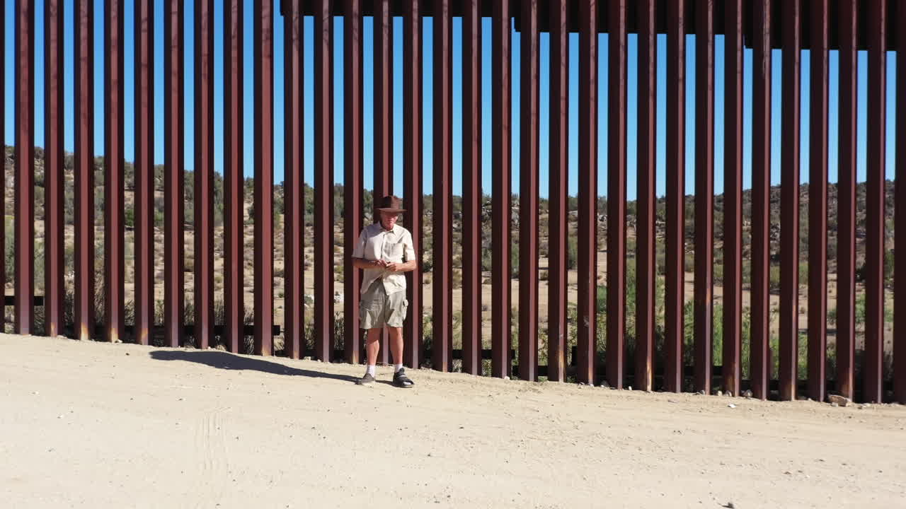 무인 항공기는 캘리포니아의 미국 멕시코 국경 벽 앞에서 모자를 쓴 나이든 미국인을 향해 날아갑니다.