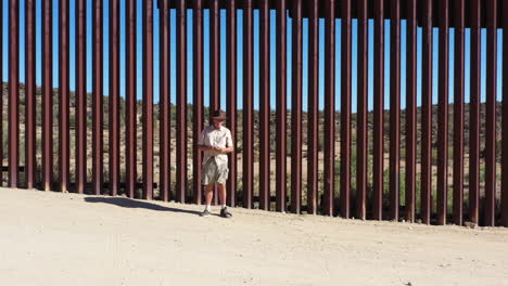 drone flies towards older american guy in hat in front of usa mexico border wall in california