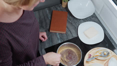 top-down view of woman stirring soup in stainless steel pot using a stick, with various cooking tools, ingredients, wooden tray, and kitchen items arranged on modern dark marble countertop