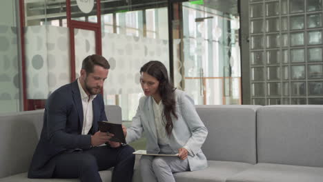 a young businessman with beard and suit has a business meeting with a pretty young businesswoman on the sofas in the common area of the office building 1