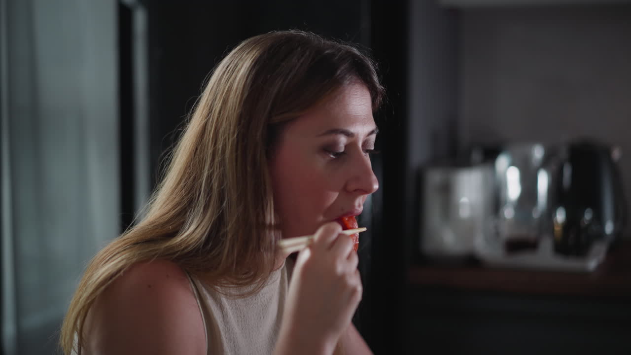 Side view of young woman eating piece of meat with chopsticks, seated indoors with softly blurred kitchen background featuring indistinct appliances