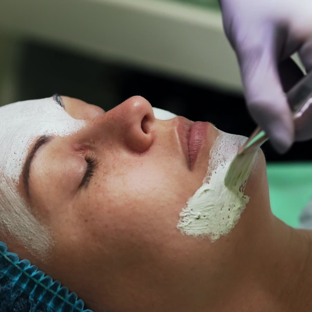 Cosmetologist covering female face with white mask. Beauty specialist uses special brush to apply the mixture. Close up
