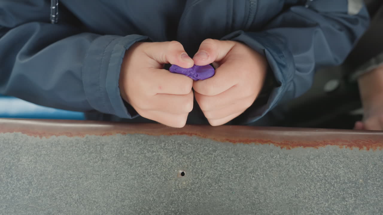 Studentenhanden kneden paarse klei alleen, close-up van geconcentreerde vingers aan een bureau met zichtbaar marineblauwe jas, rustige terug-naar-school voorbereidingssfeer, tactiele zintuiglijke oefening die fijne motoriek opbouwt, kalmte.