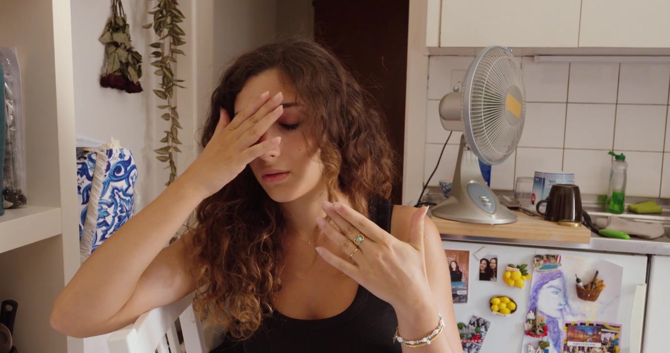 Una donna si sventola sentendosi accaldata al chiuso, mentre un ventilatore elettrico è in funzione sullo sfondo durante il caldo.